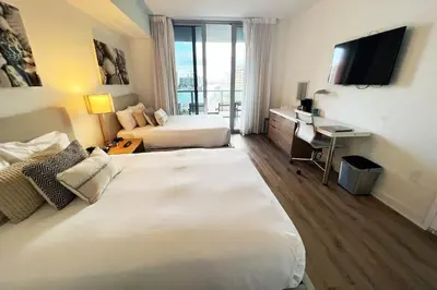 a hotel room with two beds and a desk and a television at Cozy BeachWalk Nest, 2 Full Beds, Steps to Ocean in Hallandale Beach