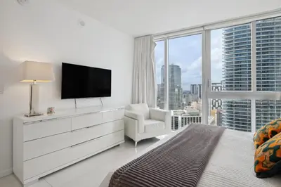a white bedroom with a tv and a chair and a window at LUX 2B-2B at ICON - Water View & 5-Star Amenities in Miami