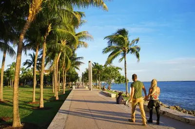 a man and woman walking down a sidewalk next to the ocean at Beachfront Bentley Hotel One Bedroom Suite with Balcony and Beach access in Miami Beach