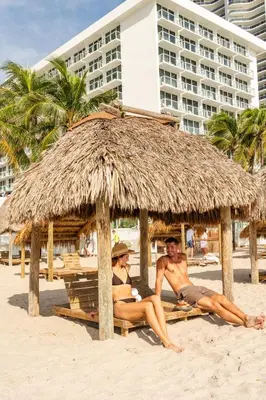 two men sitting on the beach under a thatch umbrella at The Sunny in Sunny Isles Beach